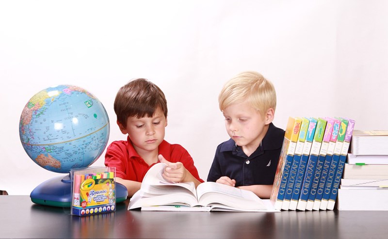 Two children sitting at a table with school supplies such as a globe, highlighters and books on it, reading a