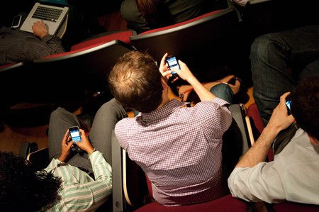 Photo from above of people sitting in a lecture hall distracted by their phones.