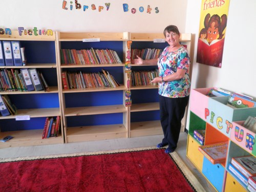 A woman poses in front of the new school library shelves which are stacked with books.