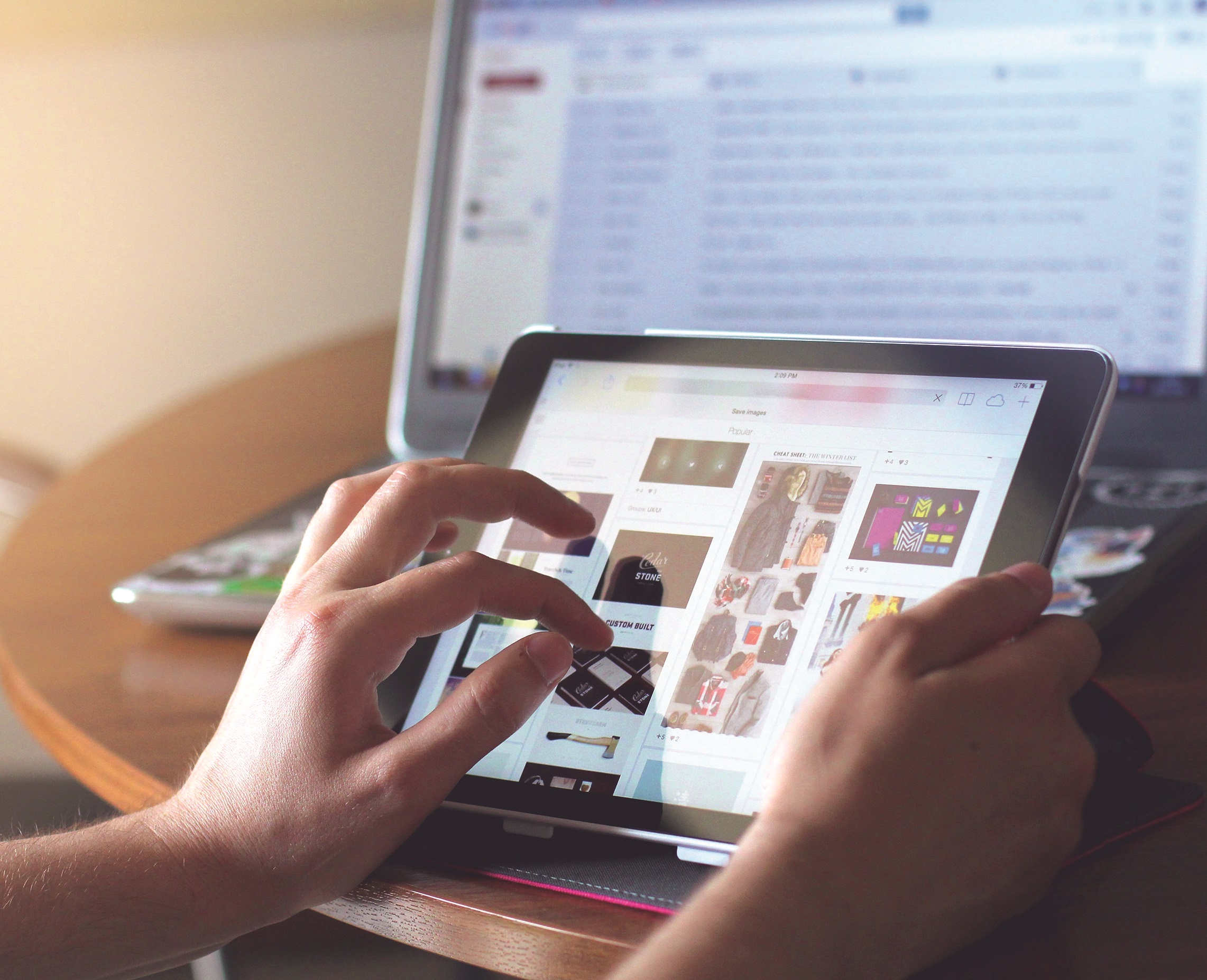 A person using a tablet device to browse images with a laptop open in the background.