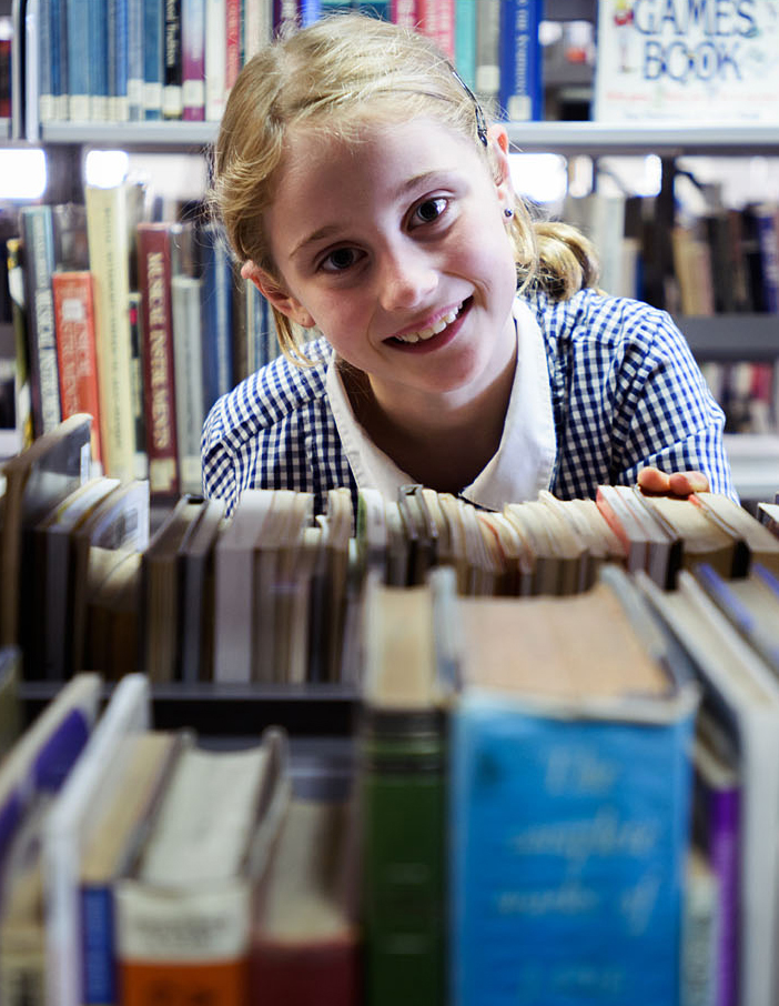 Young primary school student peering through bookshelves in a library