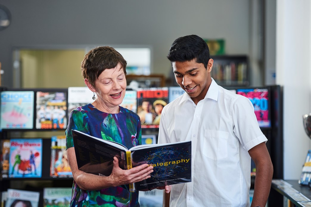 Teacher librarian with book in her hand speaking to school student