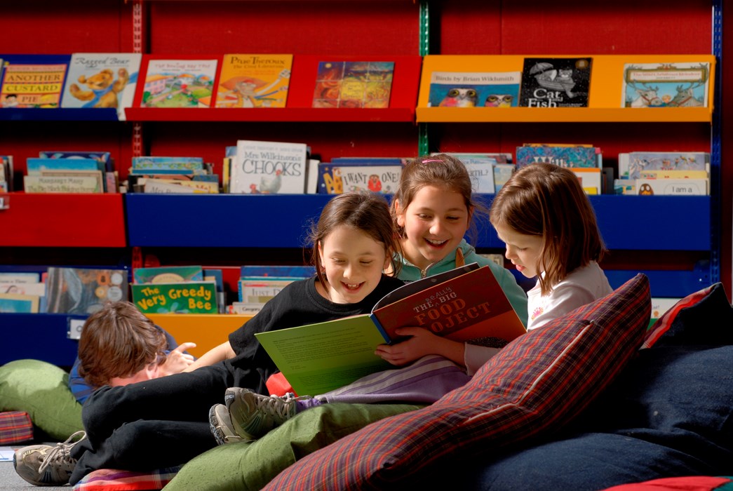 Three children in a library reading, sitting on a mound of pillows in from of book displays
