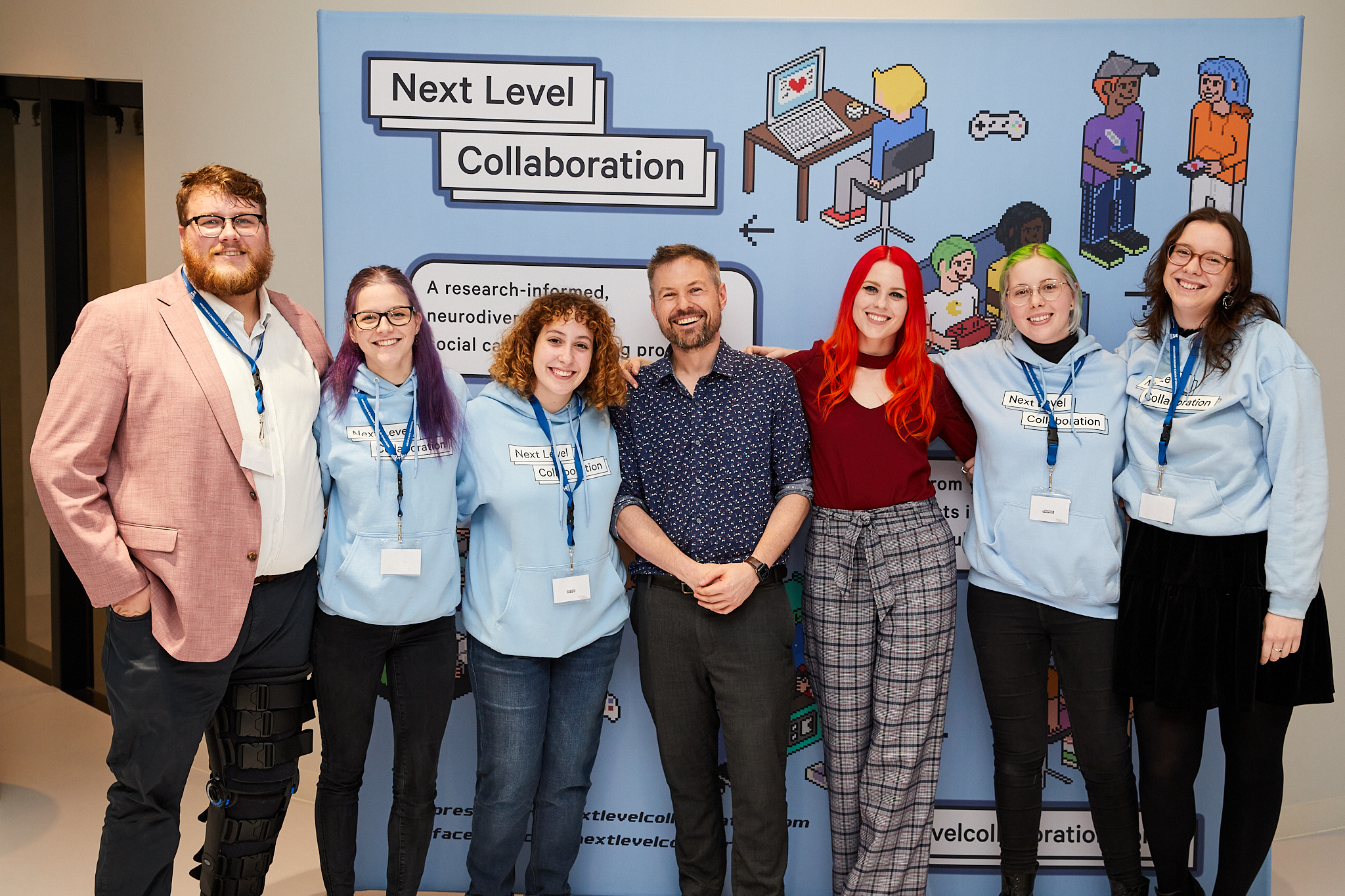 A team of seven employees from Next Level Collaboration standing in a line in front of a banner with the Next Level Collaboration logo on it.